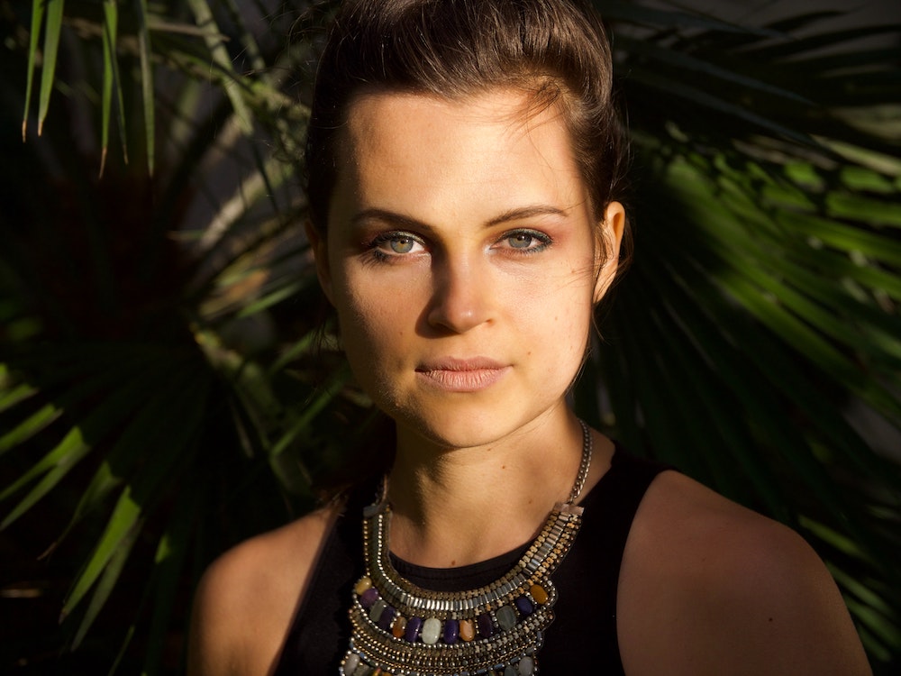 an attractive white woman wearing light makeup and statement necklace stares at the camera, against a dark background. 