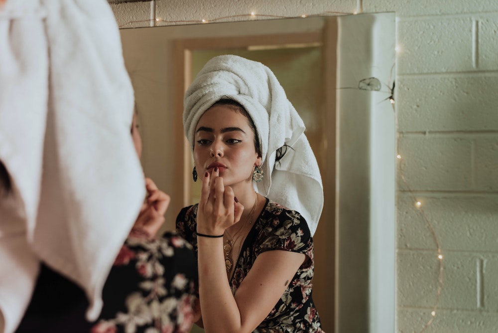 a woman with her hair wrapped in a white towel is standing in front of a bathroom mirror, applying a lip balm.