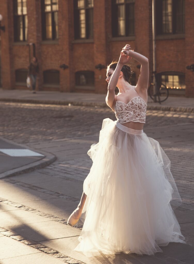 a woman in a bun, beaded bodice, and tulle skirt is doing a ballet pose in the middle of the street.