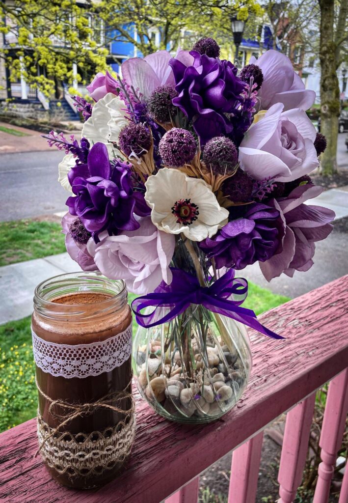 Brown_V-Whey_Protein_Shake_in_Mason_Jar_With_Brown_and_white_Lace_on_Pink_Fence_Next_to_Purple_and_White_Flowers_and_a_Green_Background_of_Trees_and_Grass