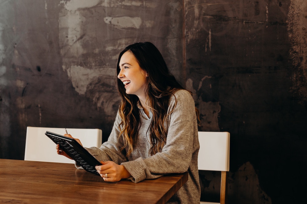 a brunette woman is sitting at a table laughing and looking at her tablet.