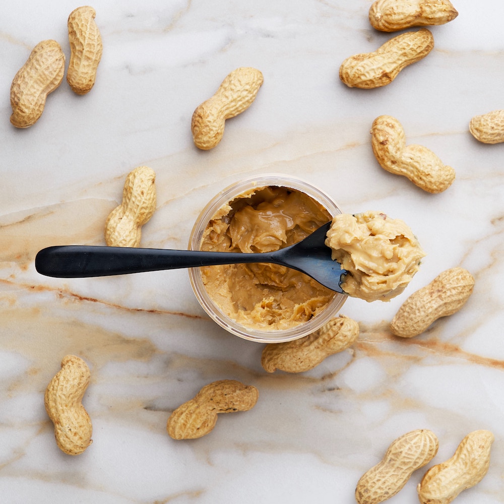 peanut butter in a jar with a spoon on top, on a marble table background. peanuts in shells are scattered around.