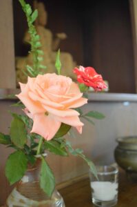 Two small roses in a ceramic vase as part of a Buddhist altar, beside an unlit candle in front of a religious figure.