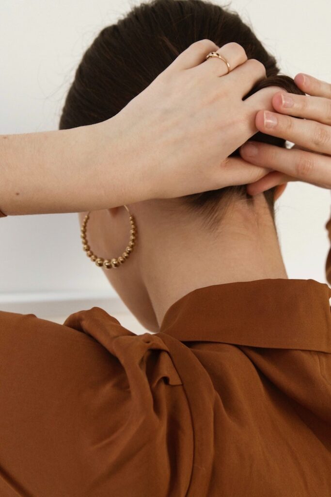 a brunette woman is shown from the back, putting her hair into a bun. she is wearing gold earrings and a sienna colored blouse. 