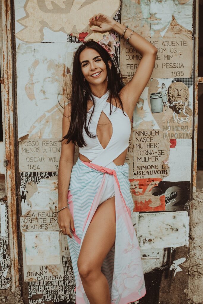 a Latin American woman in a white halter top and white skirt is leaning against a wall, smiling.