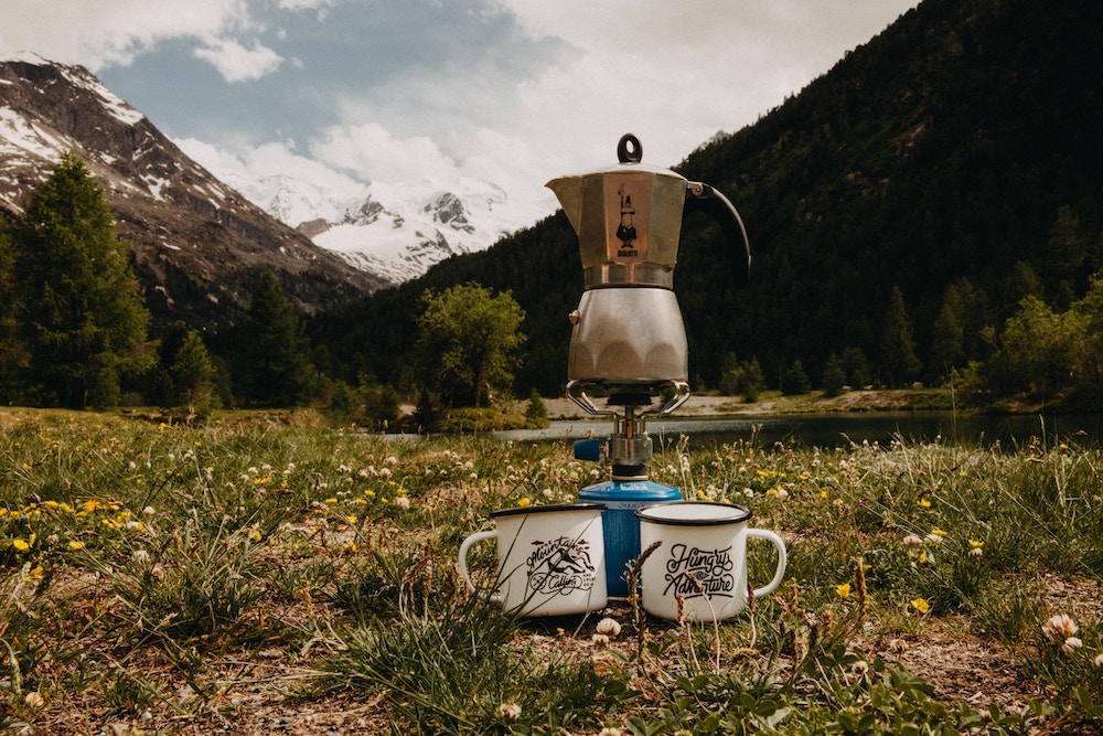 a pot of coffee on a portable burner and two mugs on a pretty green meadow with mountains in the distance.