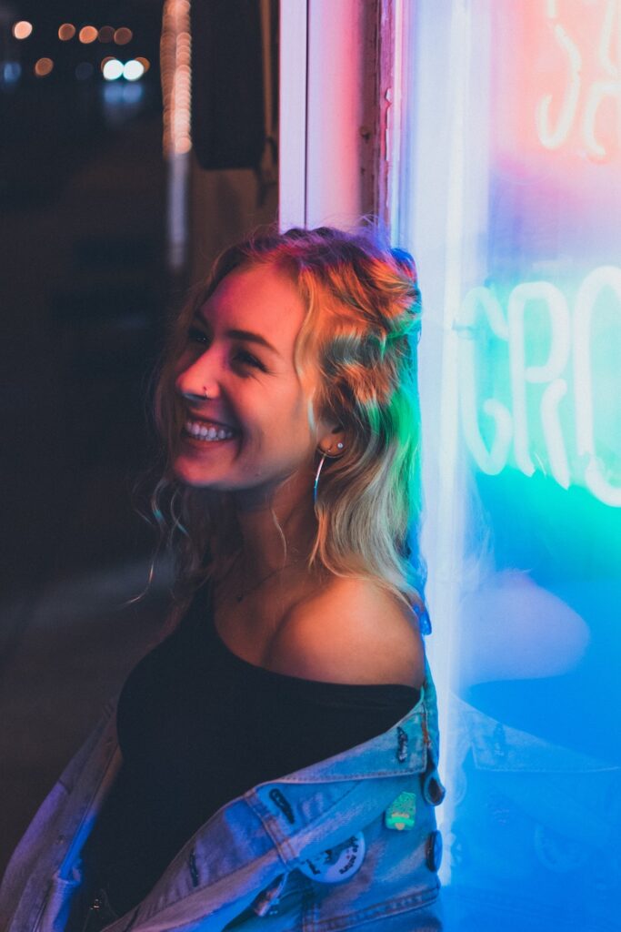 a white woman with blond hair is leaning against a neon sign, smiling.
