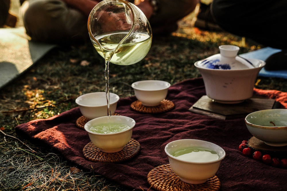 on a picnic blanket, someone is pouring clear green tea from a jug into cups.