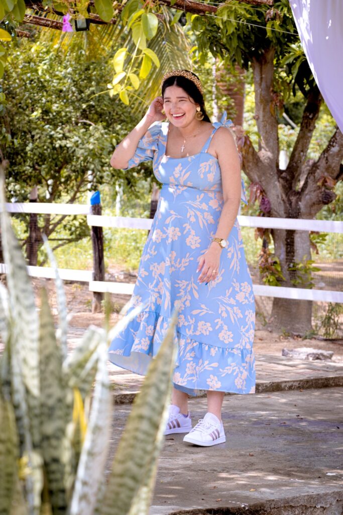 A woman wearing a light blue floral dress standing in the sun and smiling.