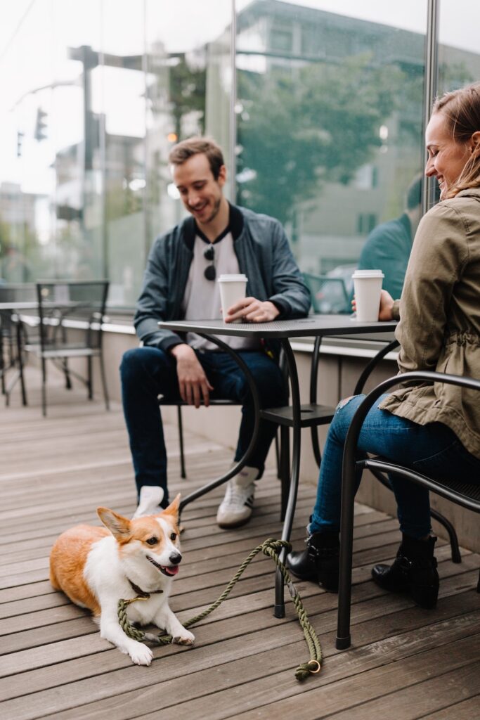 a man and a woman are sitting at an outdoor cafe table drinking coffee and smiling at their corgi.
