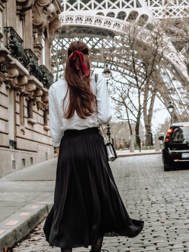 a brunette woman wearing white blouse and black skirt walking up a cobblestone street near the Eiffel Tower