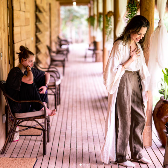 Angelina jolie stands in a white top and gray pants, smiling, in an outdoor gallery in Cambodia