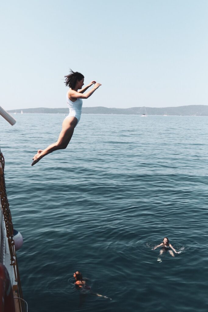 a young woman in a swimsuit is jumping off a boat into the sea as her friends look up from below. 