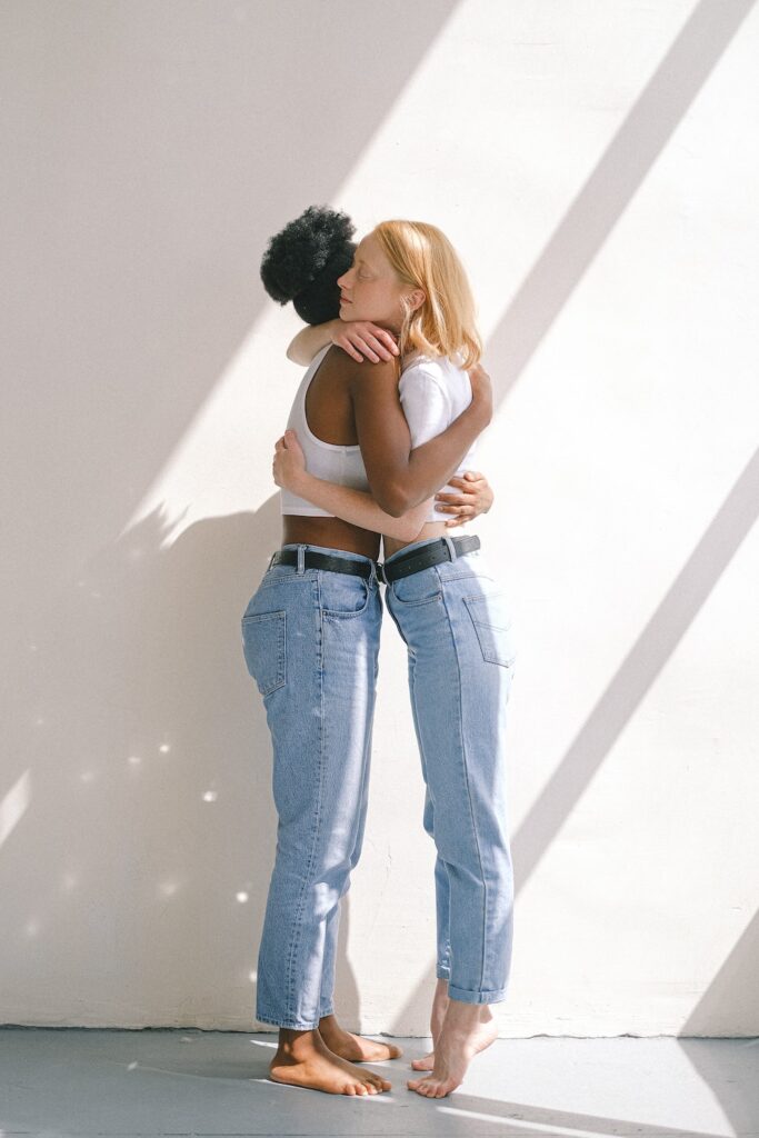 two women in white tops and blue jeans hugging in a room filled with sunlight