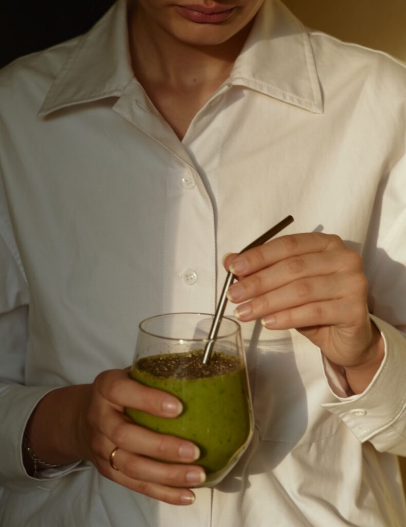 a woman in a white shirt is holding a glass of green smoothie with a stainless steel straw.
