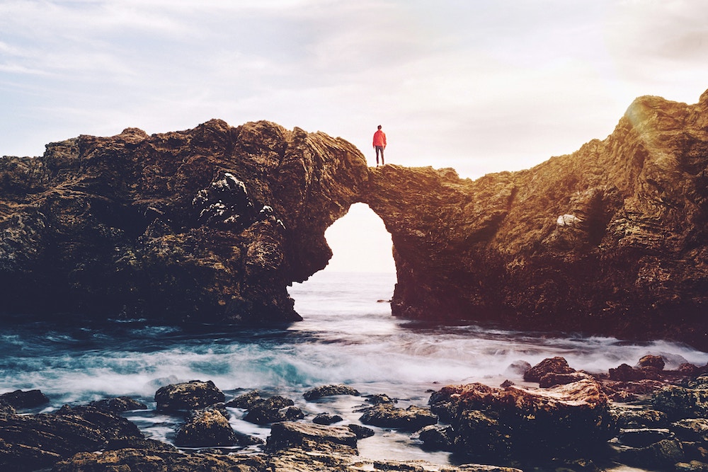 a person wearing a red jacket stands on top of a dramatic natural arch over the sea.