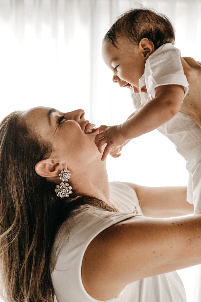 an attractive woman wearing statement earrings is raising a laughing baby high above her head.