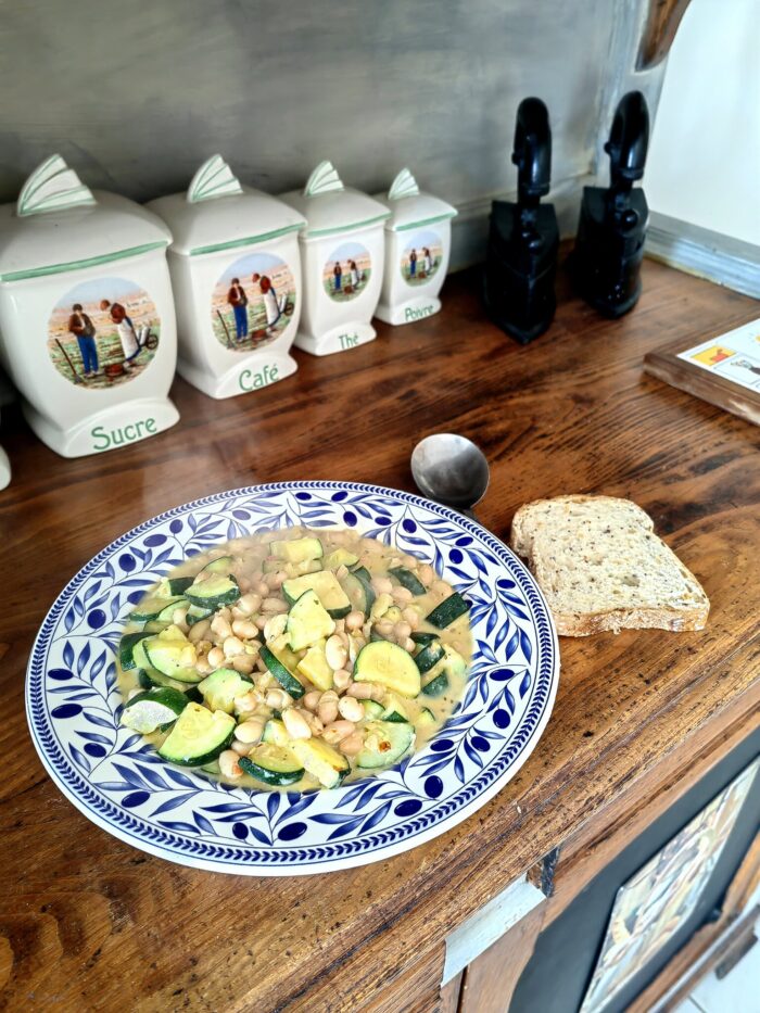 courgette in creamy garlic sauce in a blue and white floral bowl, with a side of toast, on a wooden countertop. There are various kitchen bowls and jars in the background.